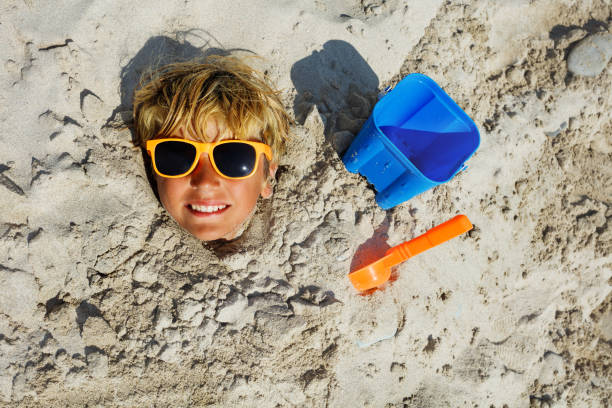 Boy's face dug into sand at the beach with all covered but the head wearing sunglasses, smile, toy bucket and scoop aside top view
