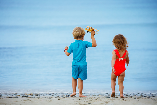 Portrait from behind of a boy with sister girl stand holding toy model of the plane over calm ocean and beach on background
