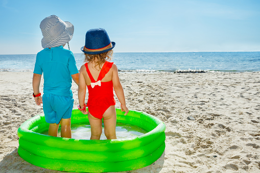 Two cute little children boy and girl, brother with sister stand in the inflatable small pool at the beach looking at the sea view from behind