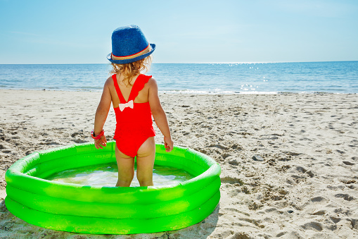Cute little girl in pink swimsuit stand in the inflatable small pool at the beach looking at the sea view from behind