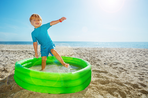 Cute blond little boy stand playing inflatable pool with big smile dancing over sea on background