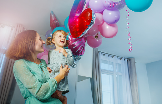 Mother holds little girl on hands with balloons decorations in living room, both smile and enjoy birthday celebration