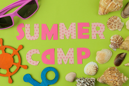 Text SUMMER CAMP of multicolored paper letters and seashells and decorative steering wheel on a bright green background. top view. flat lay