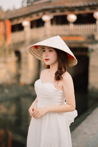 An Young vietnamese (asian) girl is standing in front of a traditional styled bridge in Hoi An. She is wearing a  nón lá (leaf hat) , which is a perfect circular cone which tapers smoothly from the base to the apex, and a white dress. It is a medium shot.