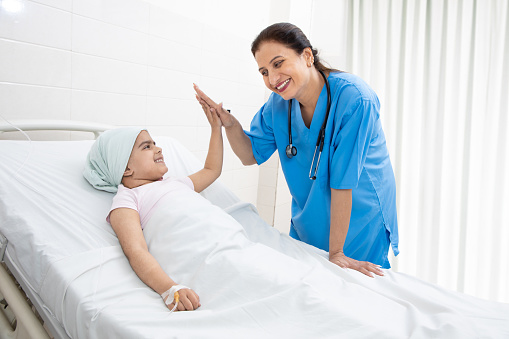 Happy indian woman nurse or medical staff give a high five to little girl cancer patient lying on hospital bed undergoing course of chemotherapy.