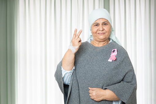 Portrait of indian senior woman cancer patient survivor standing with victory hand gesture at hospital after chemotherapy sessions , Healthcare Concept.