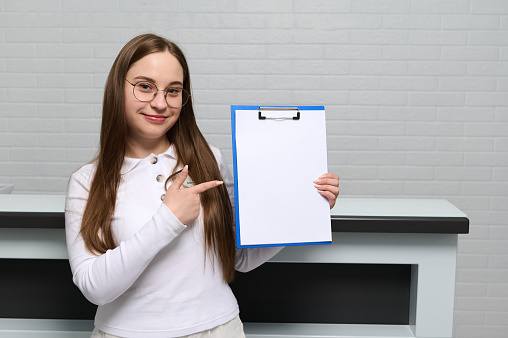 Pleasant young female receptionist at reception desk in outpatient hospital or medical clinic, showing at camera a clipboard with white blank paper sheet with free advertising space for text. Mockup