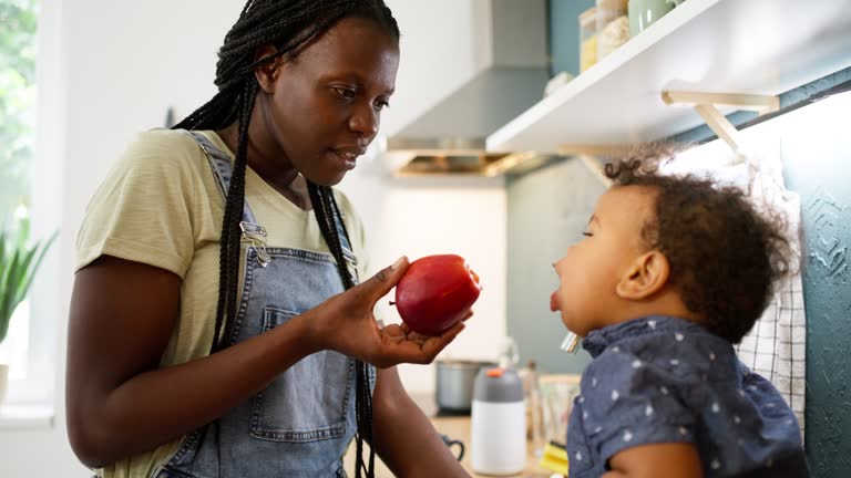 Mixed-race baby boy sitting on the kitchen counter and biting apple, while his mother having a coffee