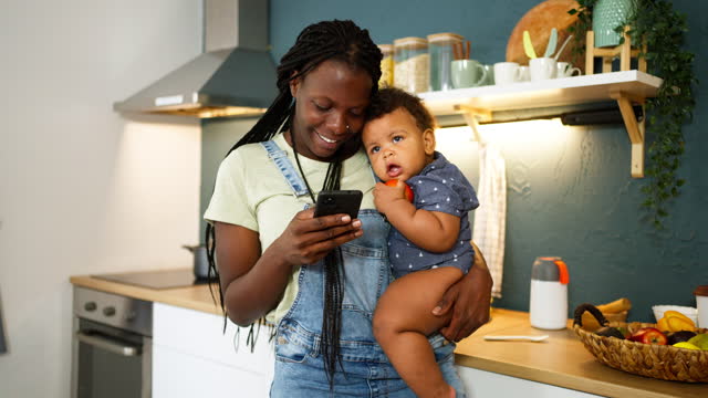 Mother Black ethnicity holding her son while using mobile phone in the kitchen