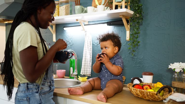 Mixed-race baby boy sitting on the kitchen counter and playing with apple, while his mother having a coffee