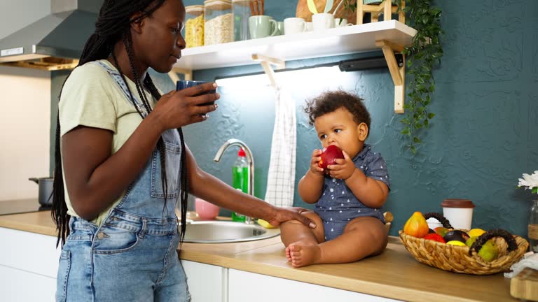 Mixed-race baby boy sitting on the kitchen counter and playing with apple, while his mother having a coffee