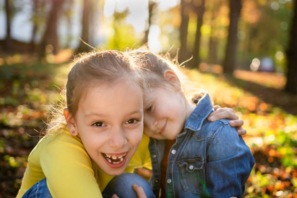Two little girls wearing summer dresses walk arm in arm along a sunny forest trail