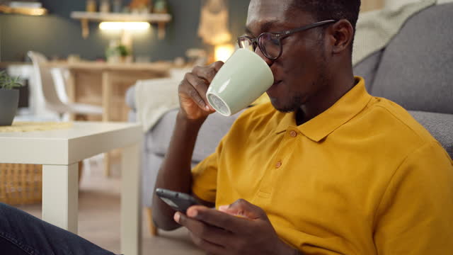 Young African-American man drinking coffee while using mobile phone at home
