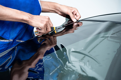 Car side window film removal and tinting installation. Male auto specialist worker hand holding cutter gently carefully cutting car front windscreen film on glass surface.
