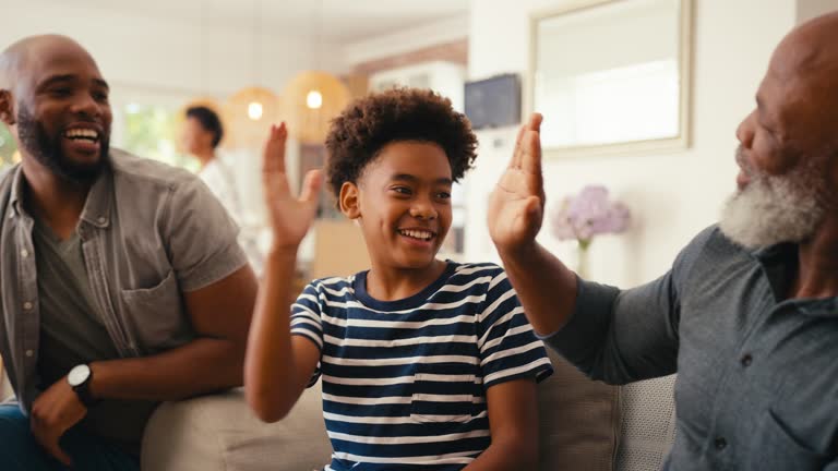 Multi-Generation Male Family Sitting On Sofa At Home Playing Video Game Together