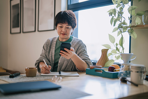 Asian Chinese mature woman reading smart phone message writing down notes at kitchen island smiling during weekend morning
