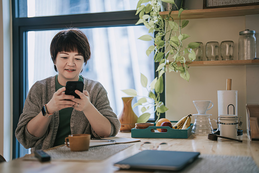 Asian Chinese mature woman reading phone text message at kitchen island smiling during weekend morning