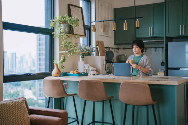 Asian mature chinese woman working at home at kitchen counter using laptop