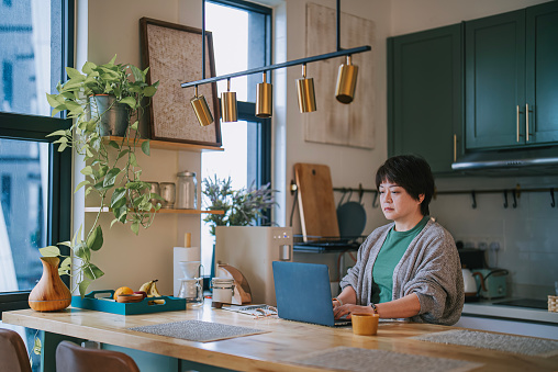 Asian mature chinese woman working at home at kitchen counter using laptop