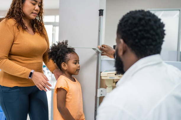 reifer arzt, der die größe schwarzafrikanischer kleiner mädchen im krankenhaus misst. attraktive professionelle medizinische männliche arbeit und medizinische untersuchung der kranken person mit familie im korridor der gesundheitsklinik. - tall human height women measuring stock-fotos und bilder