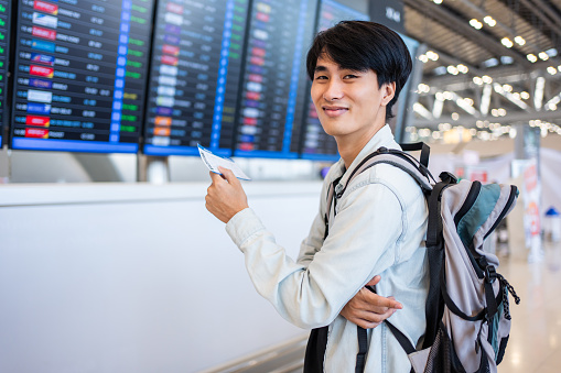 Portrait of Asian young man checking depature boarding pass in airport. Attractive male tourist passenger feel happy and excited to go travel abroad by airplane for vacation trip then look at camera.