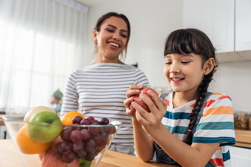Caucasian little kid biting an apple with mother in kitchen room. . Attractive mom teach and support young girl daughter take care of her body, eating a fruit after wake up for health care in house.