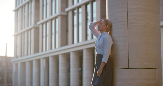 Stressed young businesswoman standing outside office. Portrait of exhausted and depressed female employee leaning against column outdoors business center