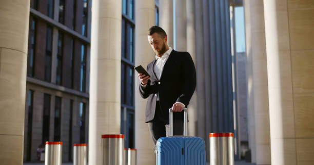istock Handsome businessman in suit standing with suitcase outside airport terminal and using mobile phone 1496054176
