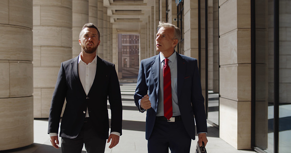 Portrait of two serious businessmen partners dressed in formal suit walking and having conversation outside business center. Confident entrepreneurs commuting to work and talking downtown