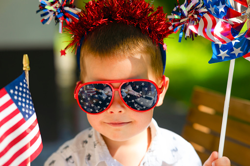 Portrait of a small child with a us flag, glasses holding jewelry and accessories. The boy celebrates Independence Day, smiles, sunny summer day, holiday, smiles. The concept of a holiday in America