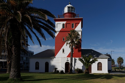 A picturesque light house is situated in a tropical environment, with lush palm trees surrounding its base and a nearby church in the background