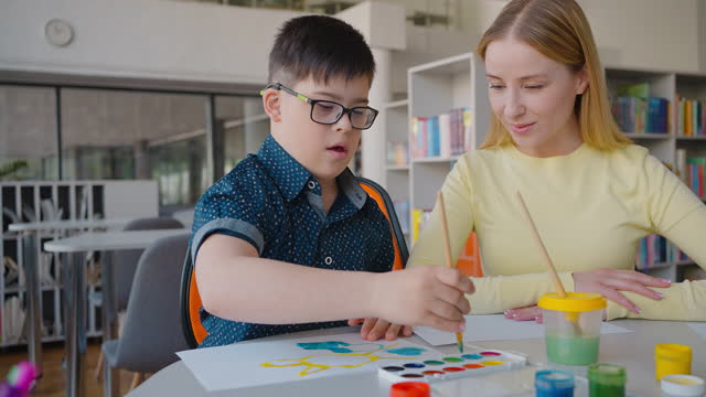 Happy elementary age boy with Down syndrome drawing watercolor picture during art class with teacher
