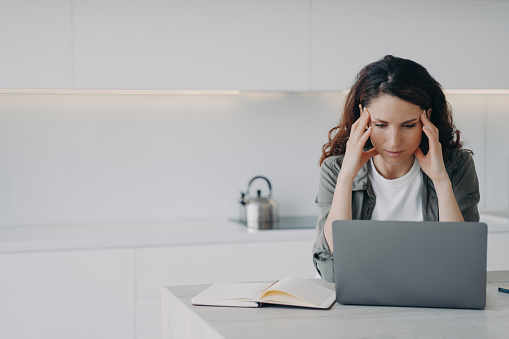 Stressed freelance worker solves laptop problems, suffers from migraine at home. Overworked woman touches temples, has headache during computer work.