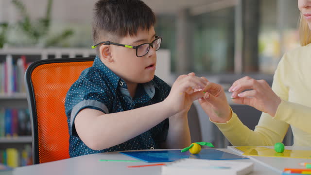 Child boy with Down Syndrome sculpts plasticine figures at art lesson under guidance of his educator