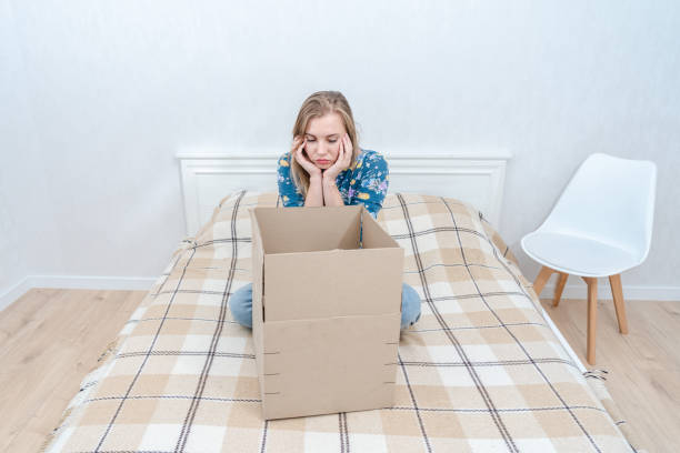 istock Sad young woman sitting on bed with open cardboard box at home. Delivering parcel 1496081587
