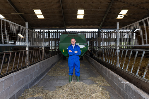 Front wide shot of a farmer standing in a barn at the sustainable farm he works at in Embleton, North East England. He is standing with his arms crossed looking at the camera.