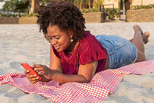Portrait of Afro woman checking her cell phone while lying on a blanket on the beach.