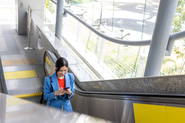 istock Asian woman using mobile phone while standing on escalator and going to office 1496087453