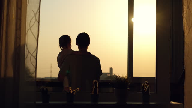 Mom holds her daughter in her arms standing at the window and launches a paper plane during sunset.