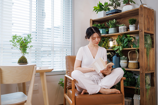 A young and beautiful Asian woman wearing white casual clothing. Sitting on the sofa in the living room, reading a book.