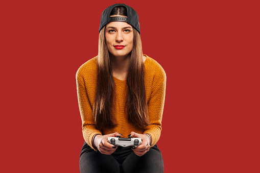 Attractive young hipster girl in snapback, sitting and holding gaming controller on red background, playing video games. Technology, gaming and lifestyle concept.