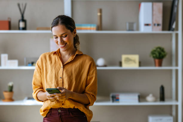 A Happy Beautiful Blonde Businesswoman Using Her Mobile Phone While Working In The Office A smiling Caucasian entrepreneur texting on her smartphone while standing indoors. happy using phone stock pictures, royalty-free photos & images
