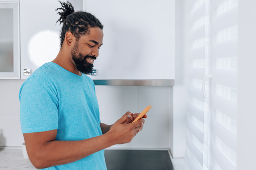 A young African American man is looking at his cellphone while standing in his kitchen.