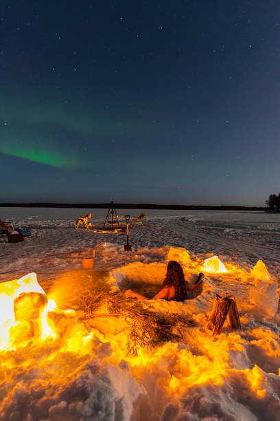 Woman enjoyng northern lights during a cold bath Rear view of a girl while ice bathing in a hole of a frozen lake under northern lights close to a campfire swedish sauna stock pictures, royalty-free photos & images