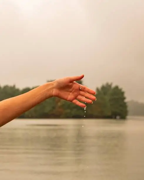 A female stands in a scenic outdoor setting with her arms outstretched, facing a tranquil lake surrounded by lush foliage
