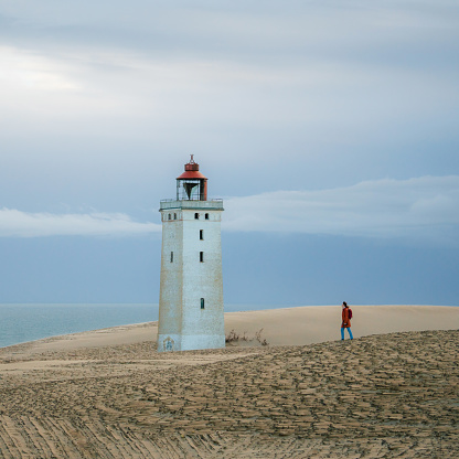 Woman in orange wind coat   walking on the background of Rubjerg Knude lighthouse during sandstorm