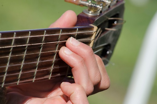 A young male musician is strumming an acoustic guitar in an outdoor setting, his hand firmly gripping the neck of the instrument