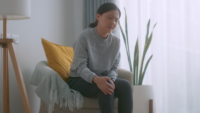 Asian woman feels pain from injury knee sitting on armchair in living room at home.