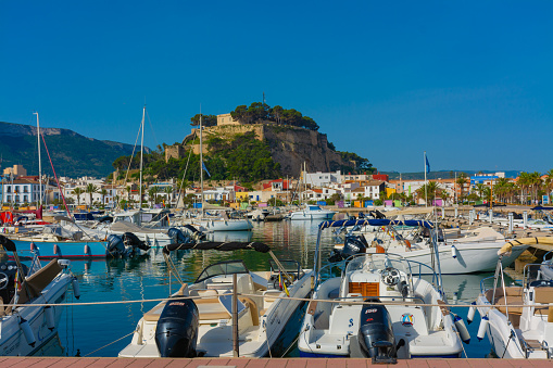 Denia, Alicante, Spain. May 22, 2022 - Sailboats and other boats moored in the marina. In the background, hotels and restaurants in the old town, and the castle on top of the hill