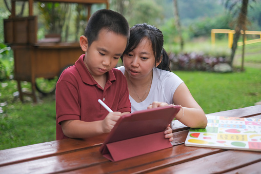 Close-up shot of Asian mother helping her son doing his preschool homework using a digital tablet. The mother and boy are sitting in a picnic table outside a restaurant terrace.